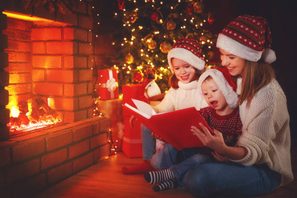 crianças e mãe de família a ler um livro no natal perto da lareira em casa - 7292 - fotografias e filmes do acervo
