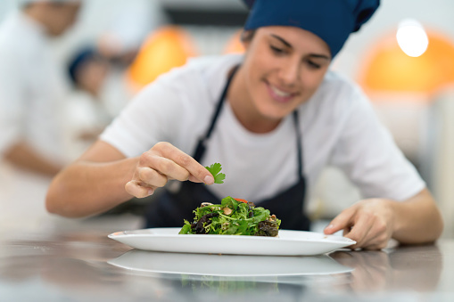 Female student at a gourmet institute finishing the details of a salad she just made looking very happy and smiling - Focus on foreground