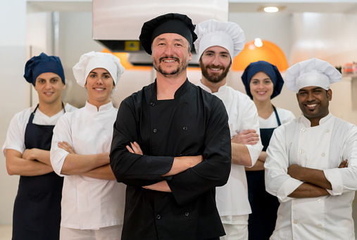 Proud multi ethnic team of chef, sous chefs and cooking assistants looking at camera smiling with arms crossed