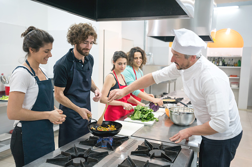 Group of people at a cooking class and chef teaching them how to sautÃ© vegetables on a wok all paying attention and smiling