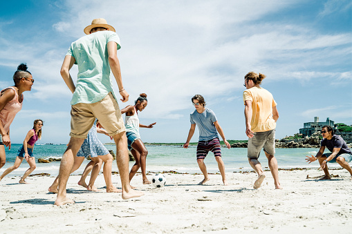 Happy multi-ethnic friends playing soccer at beach. Young males and females are in casuals. They are enjoying summer vacation.