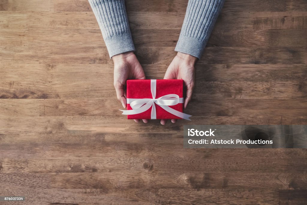 Woman hands in gray sweater on wood table giving red Christmas gift box Woman hands in gray sweater on wood table giving red Christmas gift box wrapped with white ribbon - top view with copy space Gift Stock Photo