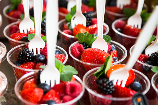 Close up high angle image depicting a display of plastic cups filled with berry fruit (strawberries, raspberries, blackberries) and sprigs of fresh mint leaves. Each cup has a white disposable plastic fork in it. Room for copy space.