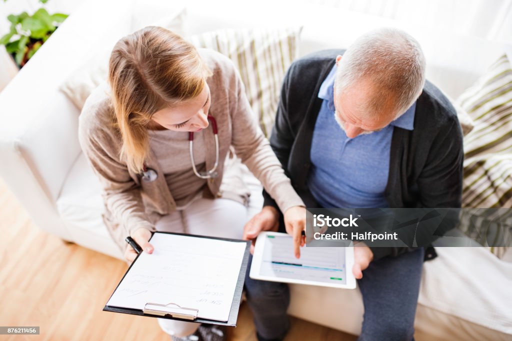 Health visitor and a senior man with tablet during home visit. Health visitor and a senior man during home visit. A female nurse or a doctor showing test results on a tablet. High angle view. Patient Stock Photo