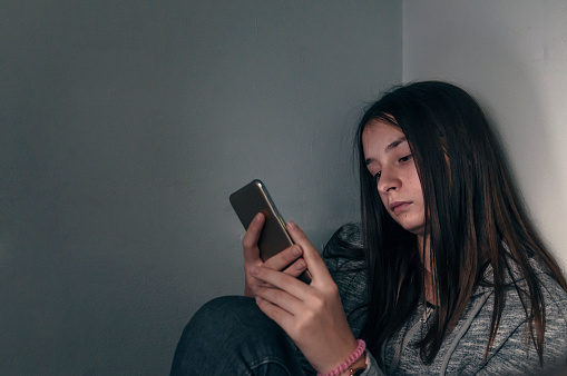 Teen girl excessively sitting at the phone at home. she is a victim of online  social networks. Sad teen checking phone sitting on the floor in the living room at home with a dark background. Victim of online bullying Stalker social networks