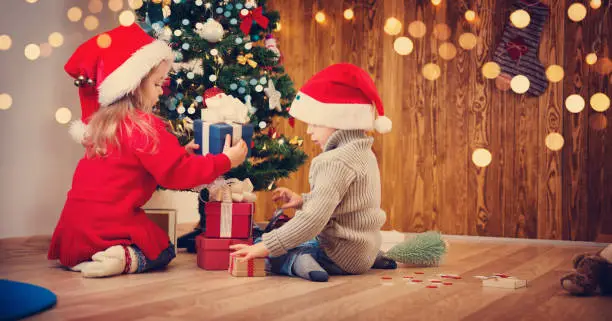 Boy and girl lying on the floor with presents near christmas tree. Children in red hats at home in winter