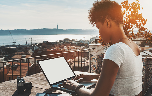 Side view of curly African American female copying recently made photos to laptop with blank mock-up screen while sitting on the balcony of Lisbon city with horizon; cityscape and river in distance