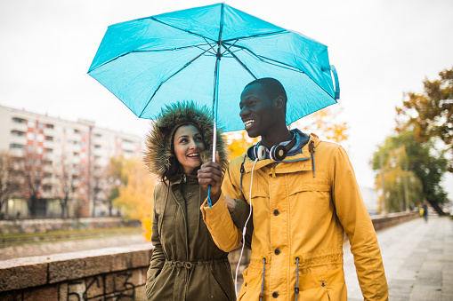 Photography of a multi-ethnic couple walking in rain and sharing an umbrella, laughing and having fun