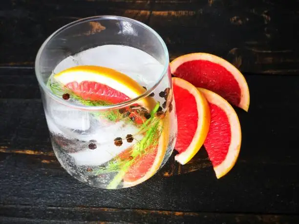 A gin and tonic cocktail garnished with a slice of grapefruit, a sliver of fennel, and black peppercorns.