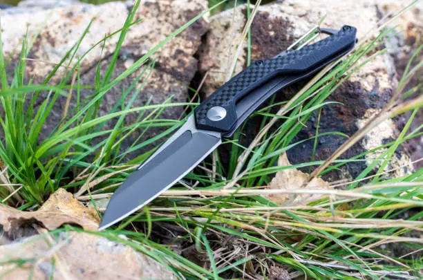 Photo of Folding knife with a black handle and a gray blade. Stones and grass.