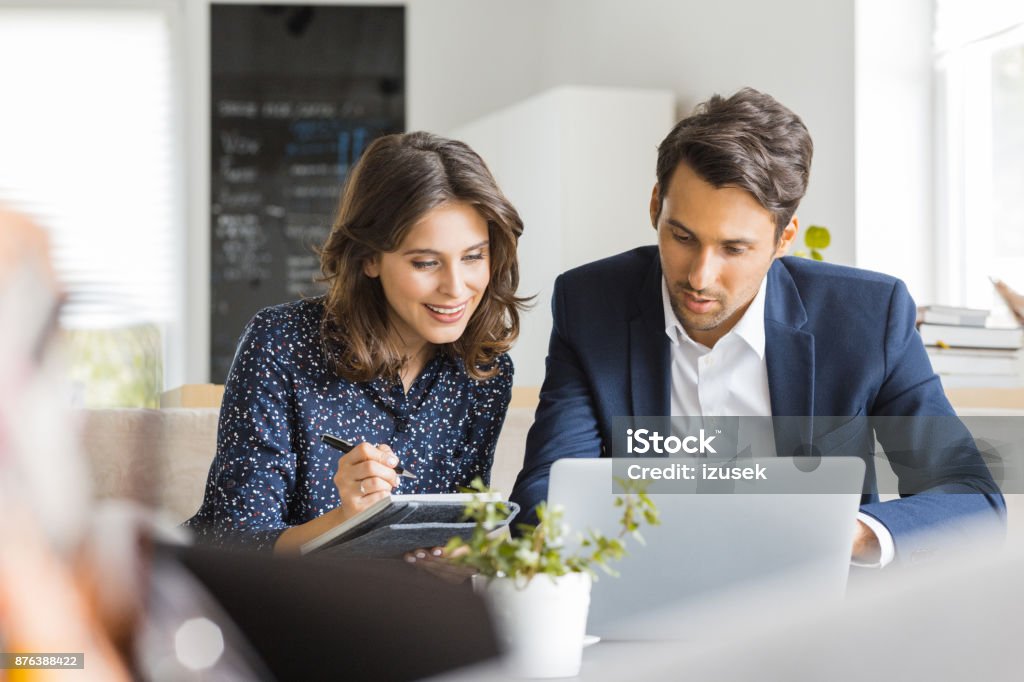 Business people working together at coffee shop Two business people sitting at cafe working on new project using laptop. Young businesswoman taking notes and businessman working on laptop computer. Advice Stock Photo