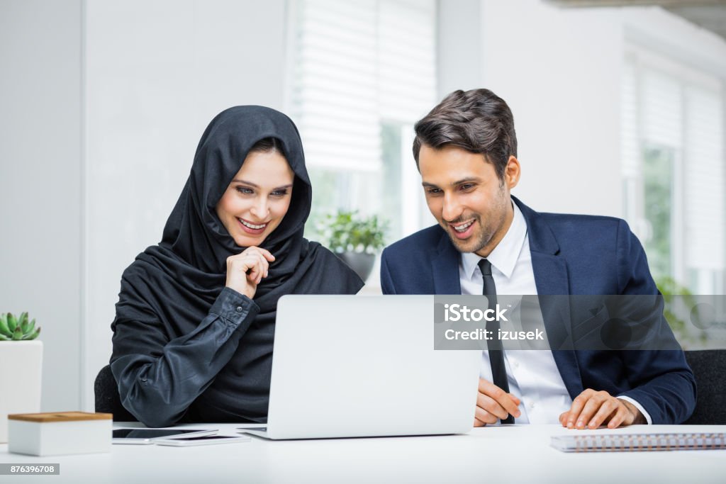 Business couple working together on project at startup office Business couple working together on project at startup office. Musilm businesswoman in hijab with male colleague looking at laptop and smiling. Coworker Stock Photo