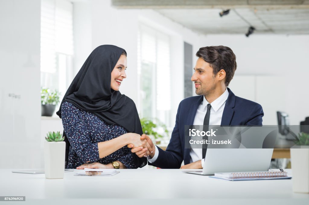Man and muslim woman shaking hands in office Two professional business people shaking hands in office. Caucasian businessman and muslim businesswoman shake hands after a meeting. Office Stock Photo