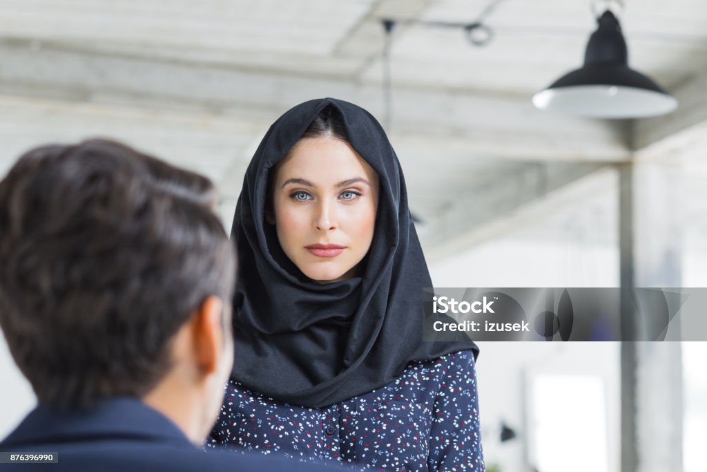 Woman with hijab looking at male colleague Woman with hijab looking at male colleague sitting in front. Businesswoman listening to businessman while working in office. Businesswoman Stock Photo