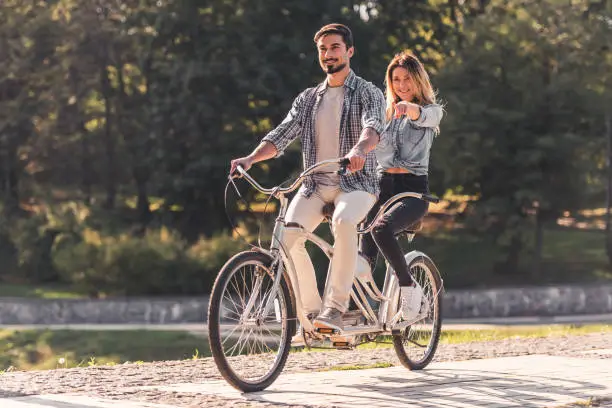Beautiful young couple is having fun while riding a tandem bicycle in the park