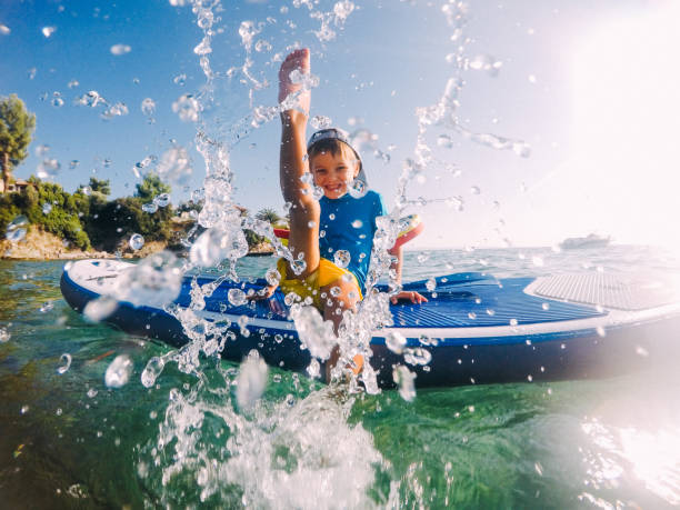 Splish-splashing Photo of a little boy being playful and splashing in the sea, while sitting on a stand-up paddle board aquatic sport stock pictures, royalty-free photos & images