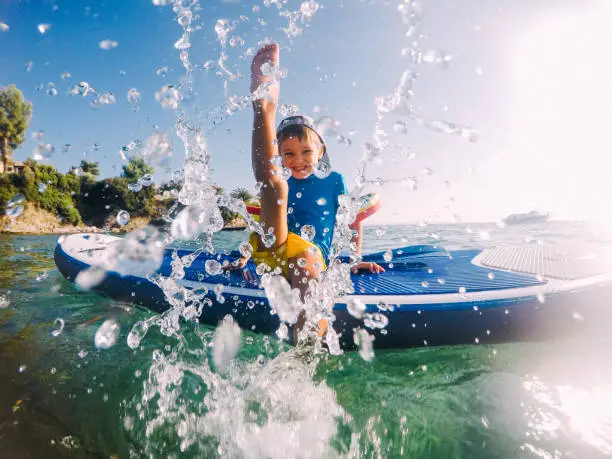 Photo of a little boy being playful and splashing in the sea, while sitting on a stand-up paddle board