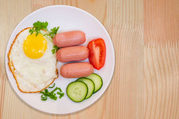 istock Breakfast fried egg and cooked mini sausages with vegetables on white round plate on light wooden table 876511494