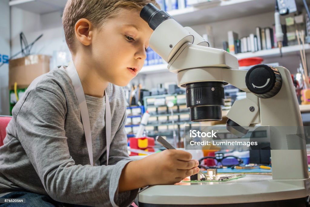 Boy repairing circuit board in IT laboratory. Little boy looking through microscope and examining mother board during science lesson. Kid repairing computer part in IT lab. Boys Stock Photo