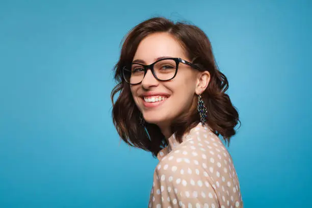 Elegant cheerful brunette in eyeglasses smiling at camera on blue background.