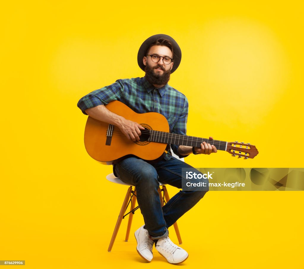 Young man with guitar in studio Content hipster man in hat and eyeglasses playing guitar sitting on chair on orange studio background. Guitar Stock Photo