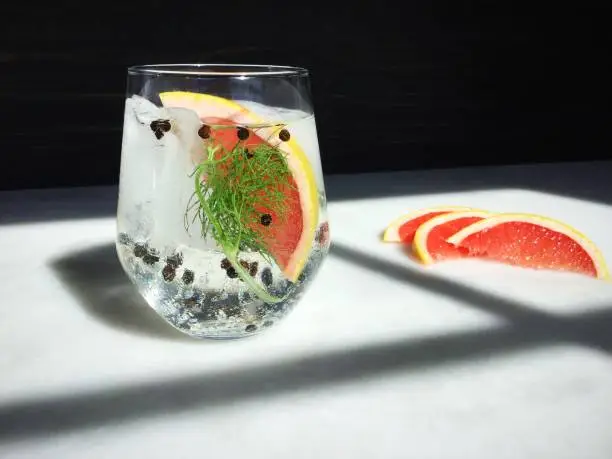 A gin and tonic cocktail garnished with a slice of grapefruit, a sliver of fennel, and black peppercorns.