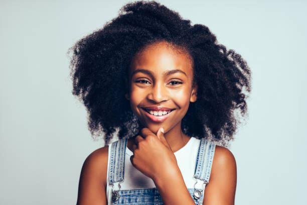 Smiling young African girl standing happily against a gray background Cute young African girl with long curly hair smiling and wearing dungarees standing with her hand on her chin against a gray background little black girl hairstyle stock pictures, royalty-free photos & images