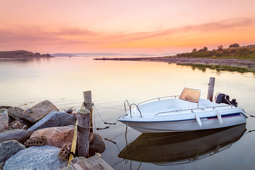 Boat, anchored at stone pier, tied to wooden  bar with rope. Dramatic morning scenery of dawn at horizon over Northern Sea in Sweden, Scandinavia, Europe. Untouched nature beauty.