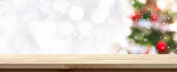 istock Wood table top Christmas holiday banner background with blurred decorated pine tree, white bokeh and snow falling in background 876774682