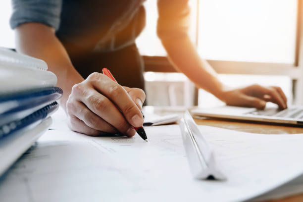 istock Young engineers holding a red pen pointing to a building on a blueprint and using a computer to write a presentation for the meeting. 876809784