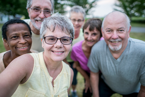 A group of senior men and women are outdoors in a park. They are taking a selfie together after doing exercise. They are wearing casual clothes.