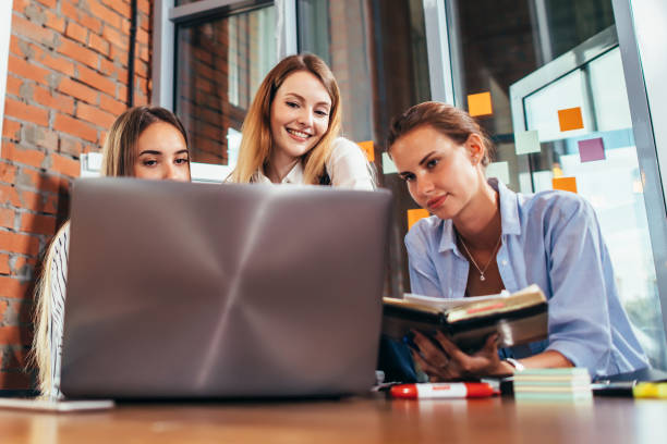 istock Three young girlfriends talking online lesson using a laptop sitting in a library 876845414