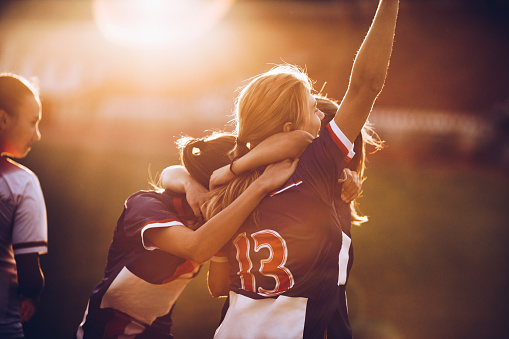 Team of happy female soccer players celebrating their achievement on a playing field at sunset.