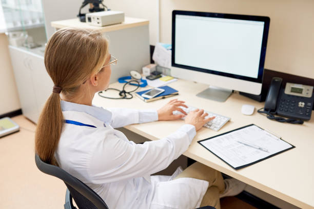 Female Doctor Working at Desk Back view portrait of blonde female doctor sitting at desk in office working with computer, typing and looking at blank white screen, copy space medical examination room stock pictures, royalty-free photos & images