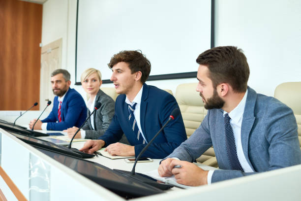 Young Politician Speaking in Press Conference Portrait of several business people sitting in row participating in political debate during press conference answering media questions speaking to microphone media interview photos stock pictures, royalty-free photos & images