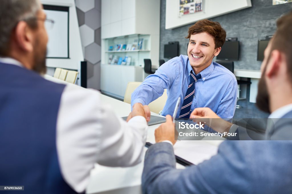 Young Businessman in Greeting Partners in Meeting Portrait of smiling young businessman shaking hands with partners at meeting table in board room greeting each other before negotiations Job Interview Stock Photo