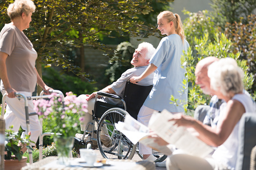 Nurse supporting senior man in wheelchair during meeting with friends in garden on sunny day