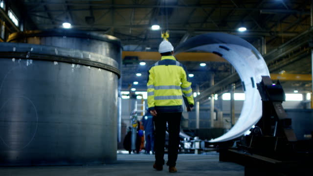 Back View of Industrial Engineer in Hard Hat Wearing Safety Jacket Walks Through Heavy Industry Manufacturing Factory with Various Metalworking Processes.