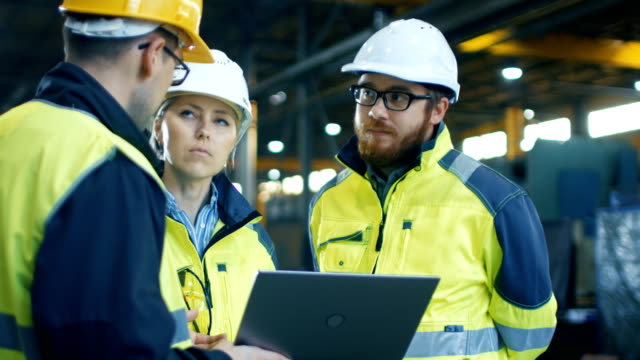 Male and Female Industrial Engineers Talk with Factory Worker while Using Laptop. They Work at the Heavy Industry Manufacturing Facility.