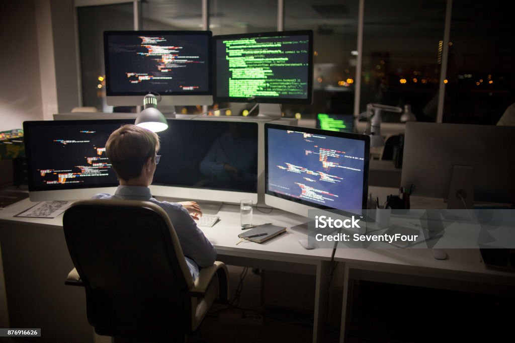 Talented Coder at Workplace Back view of highly professional coder looking at wristwatch while sitting in front of computer and working on ambitious project, interior of dim open plan office on background Computer Hacker Stock Photo