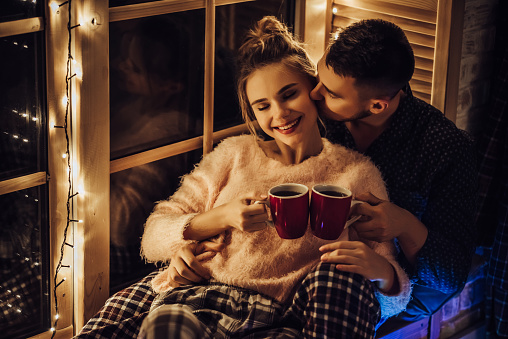 Young romantic couple is spending time together at home in pajamas.Sitting on window sill with garland in New Year Eve.Waiting for Christmas.Two lovers are hugging and kissing in Saint Valentine's Day