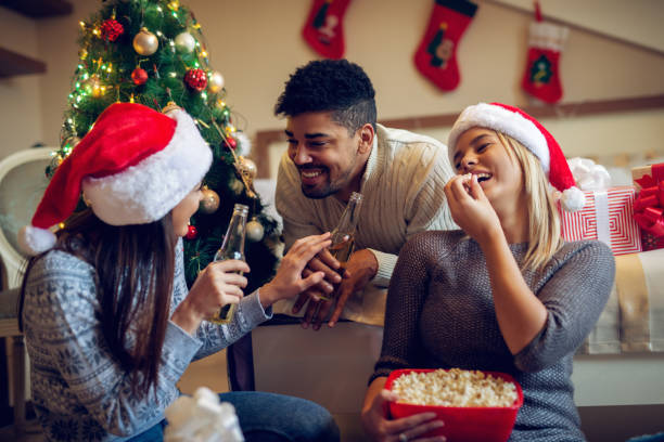 istock Three attractive stylish friends drinking beer and eating popcorn while talking funny things and sitting near Christmas tree in the bedroom. 876971750