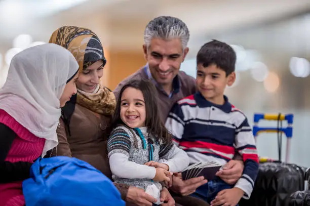 A Middle-eastern father, mother, brother and two sisters have just arrived to a new country. They are happily sitting together and smiling. The father is holding his family's passports. Their luggage is sitting to the side.