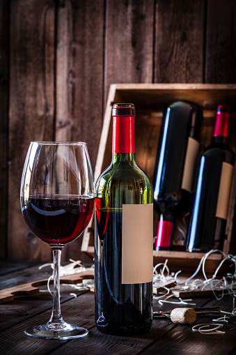 Vertical view of a wineglass and a red wine bottle shot on rustic wooden table. At the background and out of focus is a wooden box with two wine bottles. Predominant color is brown. Low key DSRL studio photo taken with Canon EOS 5D Mk II and Canon EF 100mm f/2.8L Macro IS USM