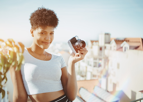 Dainty young cheerful African American female with film retro camera on the roof; smiling black girl holding vintage photo camera while standing on the balcony of house with cityscape in background