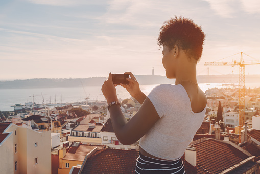 Side view of curly African American female standing on the roof of house and taking panoramic shoot on her smartphone; sunny day, Lisbon city with Tagus river and residential area in background