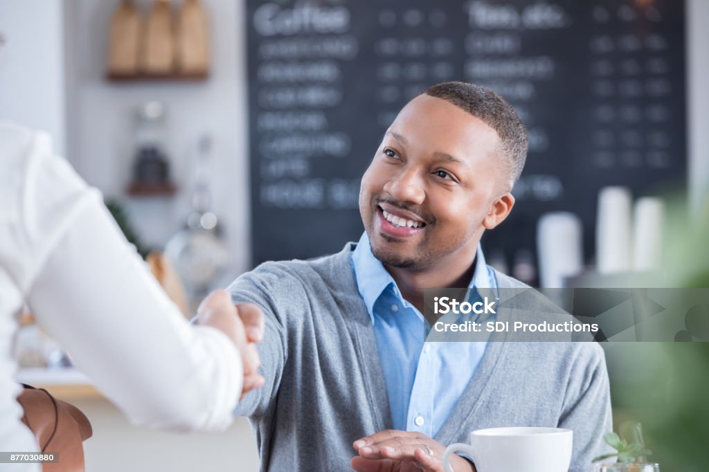Businessman meets interviewee in coffee shop A handsome mid adult businessman sits at a table in a coffee shop and shakes hands with an unrecognizable interviewee.  They are meeting for the first time as he gestures for her to sit down. Interview - Event Stock Photo