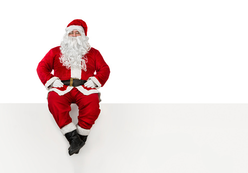 Santa Claus sitting at the edge of a blank banner isolated on white background with copy space