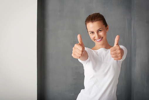 Shot of a successful young woman giving thumbs up while standing at grey wall.