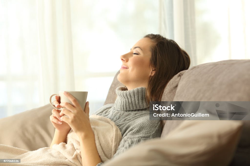 Woman relaxing at home holding a coffee mug Woman relaxing holding a coffee mug sitting on a sofa in the living room in a house interior Winter Stock Photo